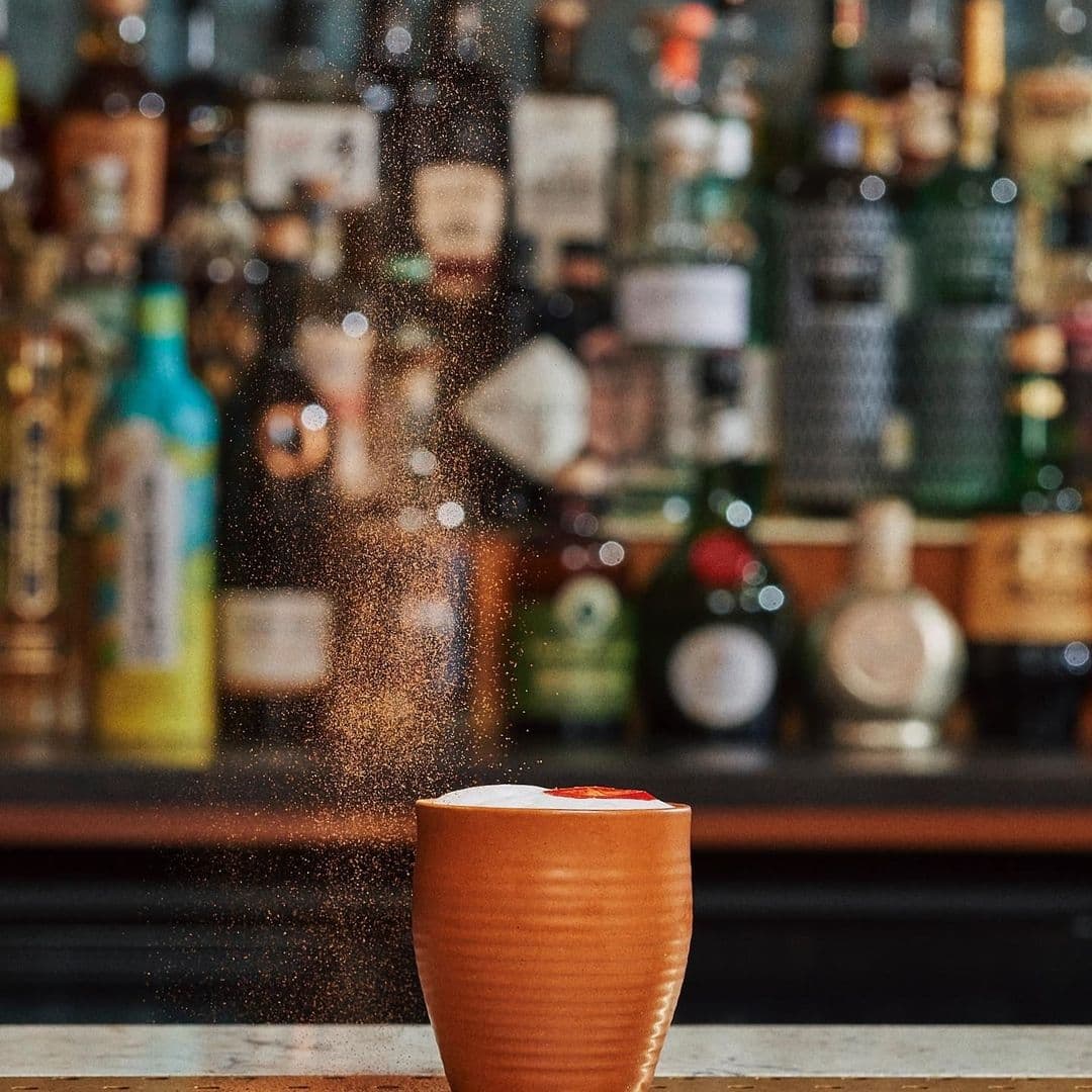 A crafted cocktail in a terracotta cup garnished with a citrus slice, with a dynamic sprinkle of spices against the backdrop of the well-stocked bar at Farrands Bar.