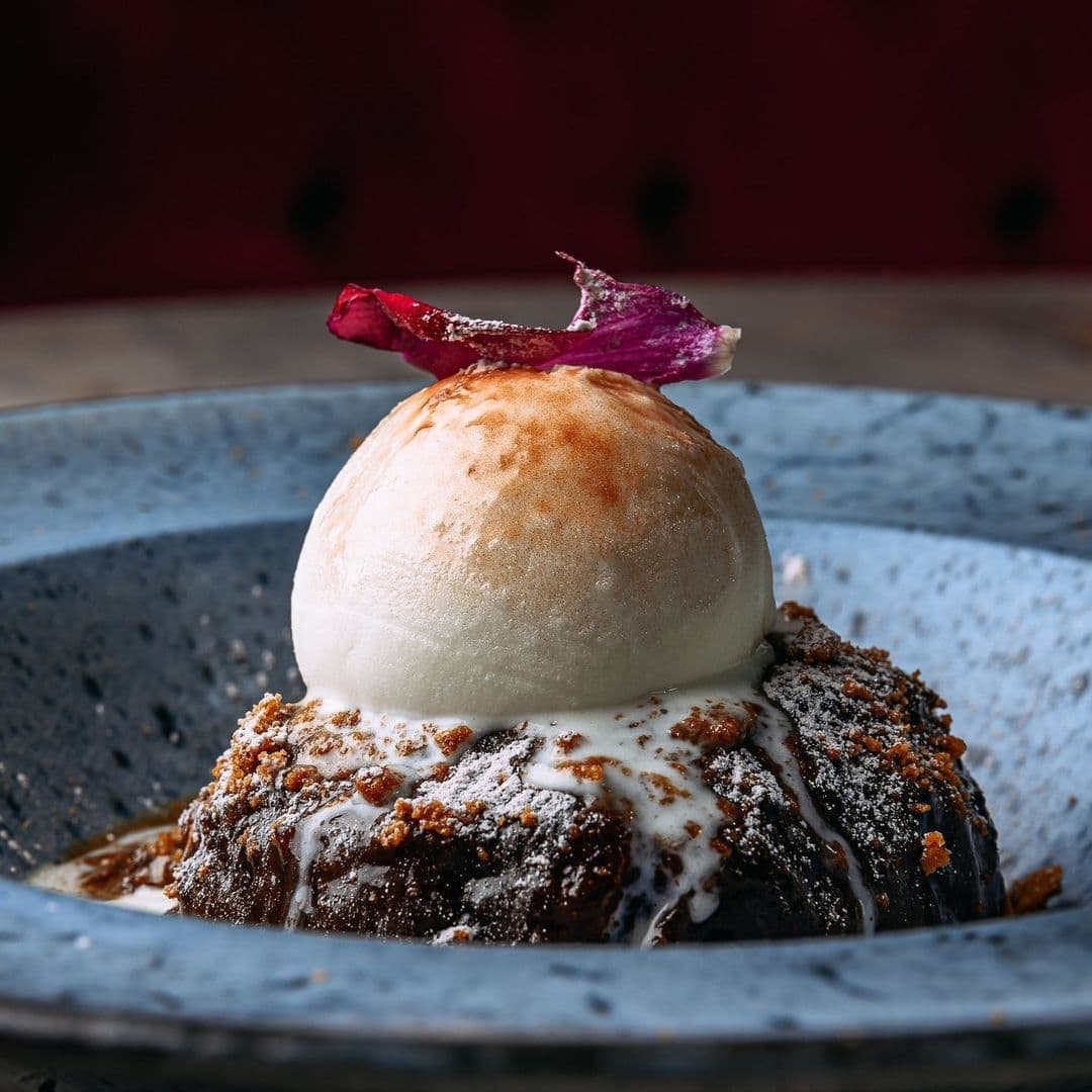 Decadent dessert at Fleur Restaurant and Bar featuring a scoop of ice cream on top of a moist, dark cake, garnished with a pink flower petal, served on a textured blue plate.