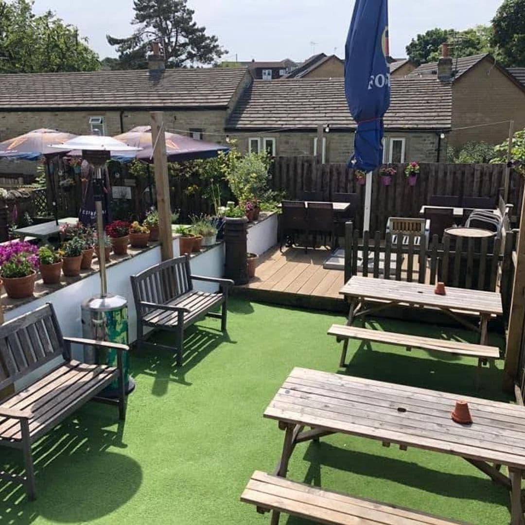 Sunny outdoor seating area at Fox & Grapes with wooden benches, umbrellas, and potted plants on artificial grass.