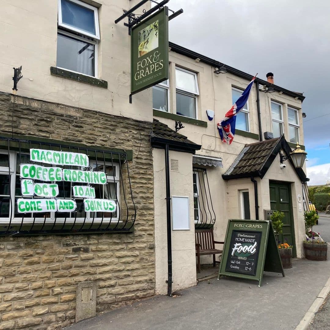 Exterior view of the Fox & Grapes pub with a stone facade, displaying a hanging sign with the establishment's name and a British flag. A neon sign announces an event, and a chalkboard advertises homemade food with serving times.