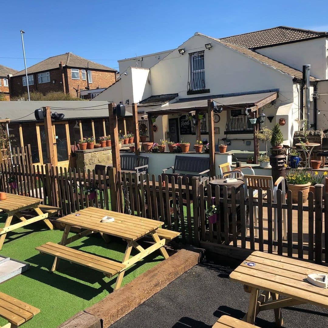 Sunny outdoor seating area at Fox & Grapes with wooden picnic tables, green artificial turf, and decorative potted plants.
