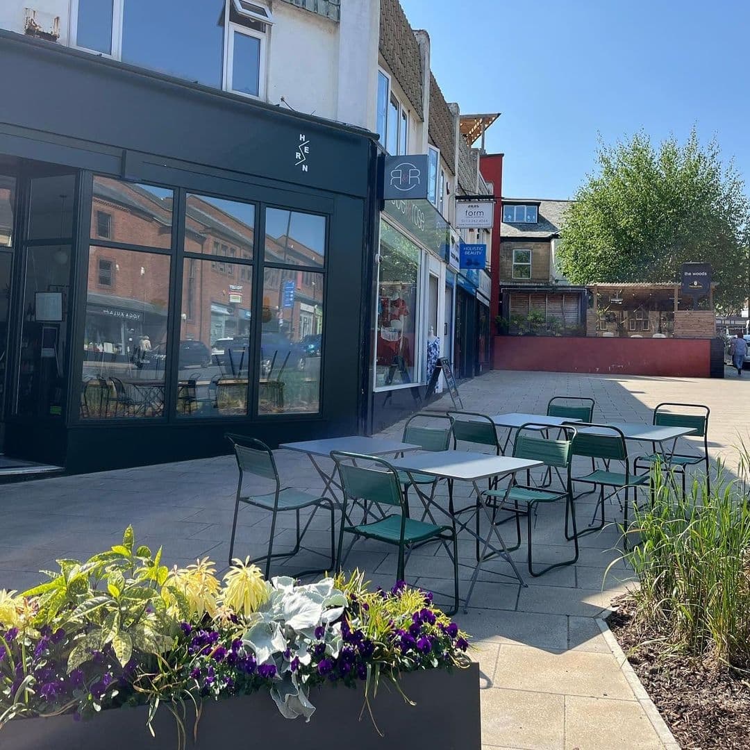 Outdoor seating area with green metal chairs and tables in front of a modern black storefront at Hern, under a clear blue sky. Vibrant flower planters add a touch of nature to the urban setting.