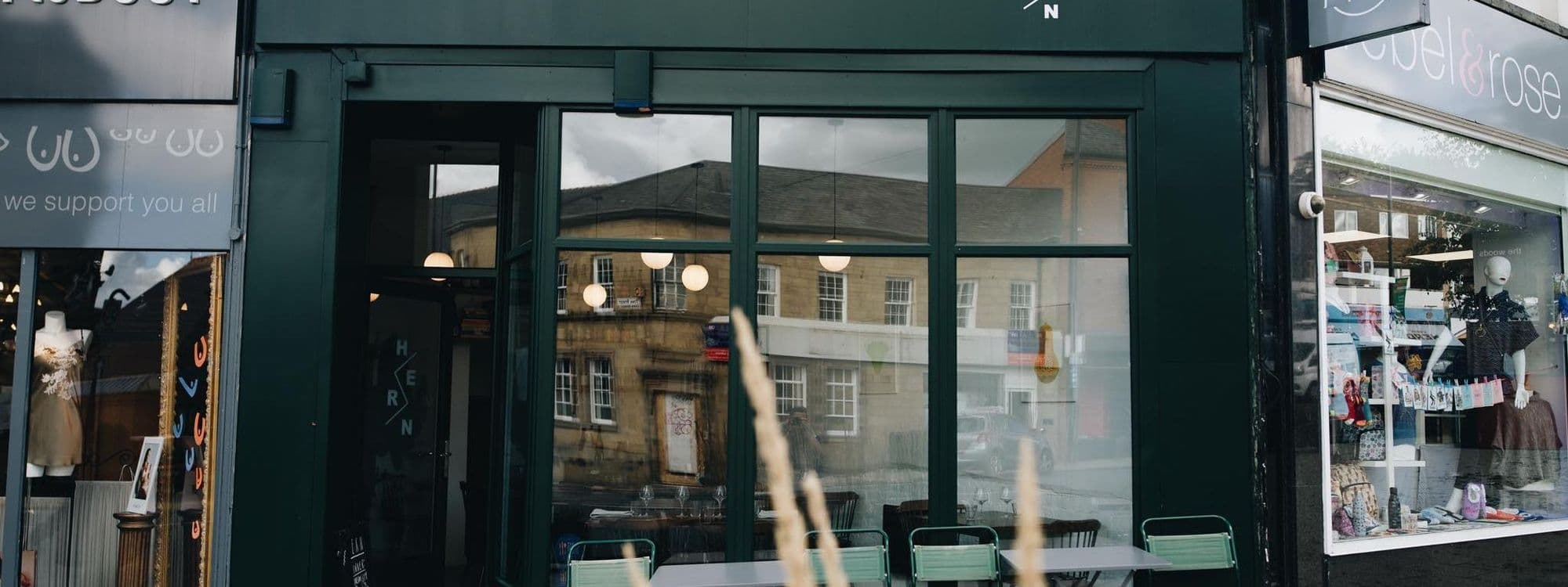 Exterior view of Hern cafe with reflective windows showing the street and buildings opposite, and a neighboring shop with displayed items.
