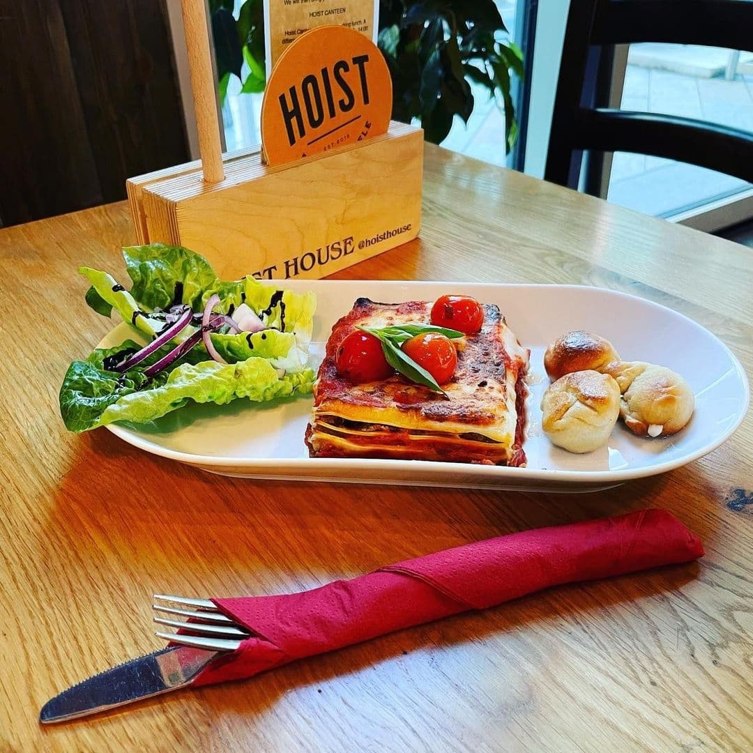 Delicious lasagna with cherry tomatoes on top, served with a side salad and bread rolls on a white plate at Hoist House.