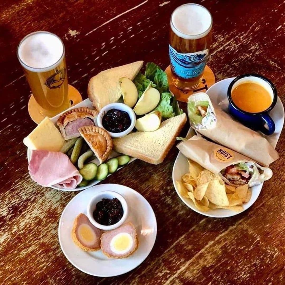 Ploughman's lunch with assorted cheeses, ham, pickles, and bread, Scotch egg on a plate, wrapped sandwiches, crisps, and two pints of beer on a wooden table at Hoist House.