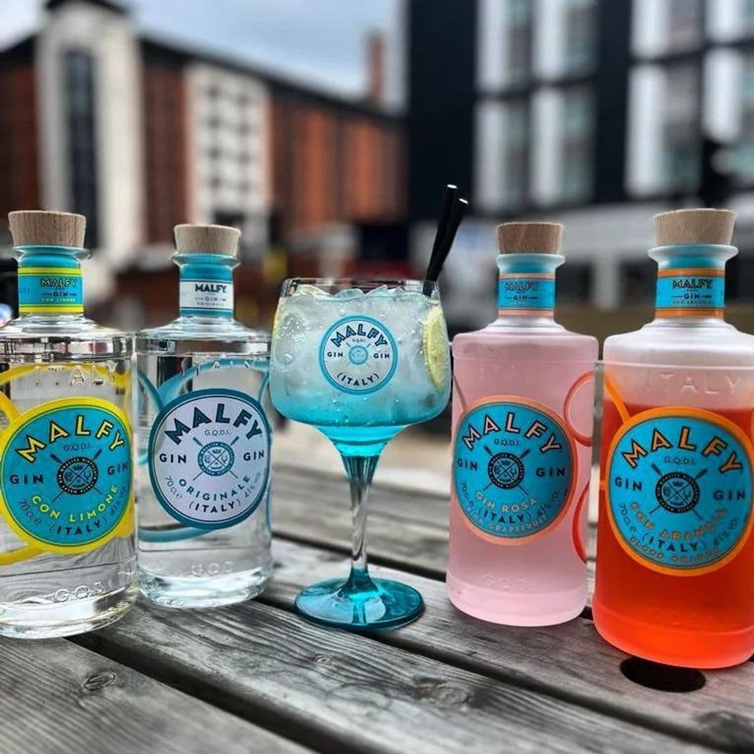 Assorted Malfy gin bottles and a cocktail glass on a wooden table at Hoist House with blurred building background.