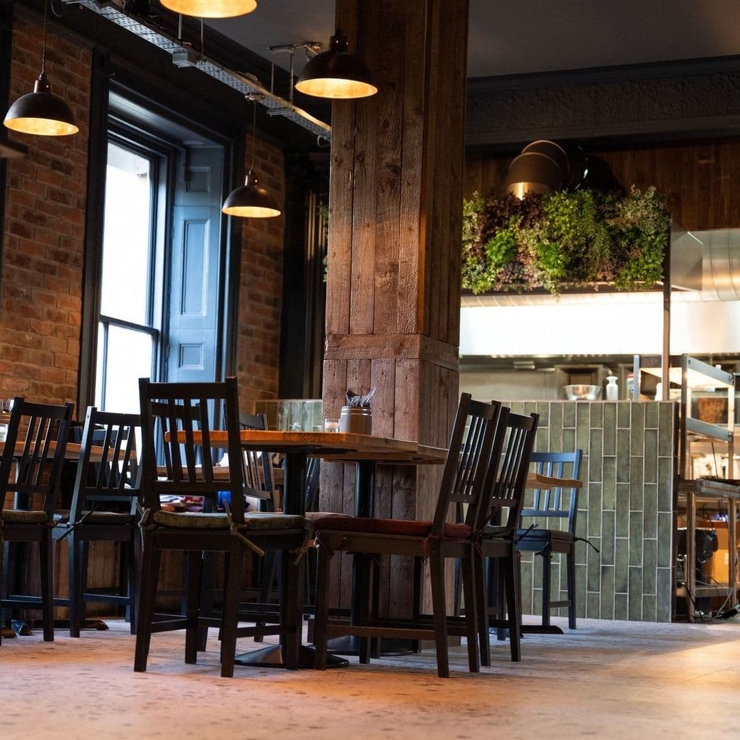 Interior of MYTHOS Chapel Allerton showing wooden tables and chairs, exposed brick walls, pendant lighting, and a visible kitchen area with greenery.