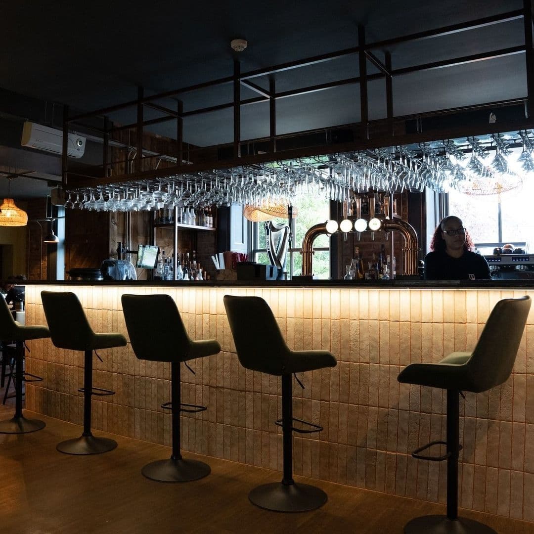 Interior of MYTHOS Chapel Allerton showing a stylish bar with hanging wine glasses, a row of high stools, and a bartender behind the counter.