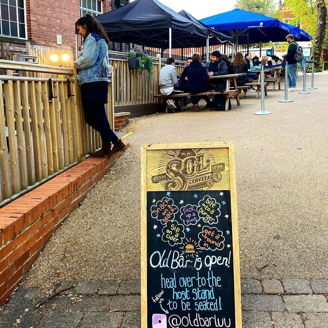 Outdoor seating area at Old Bar – Leeds Uni with people socializing, a sandwich board announcing the bar is open, and a woman standing by the railing.