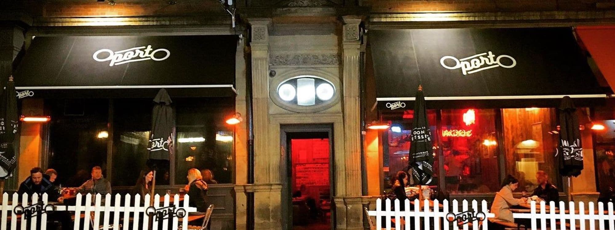 Evening ambiance at a dining venue with patrons seated under awnings labeled "Oporto," warm lighting, and a white picket fence.