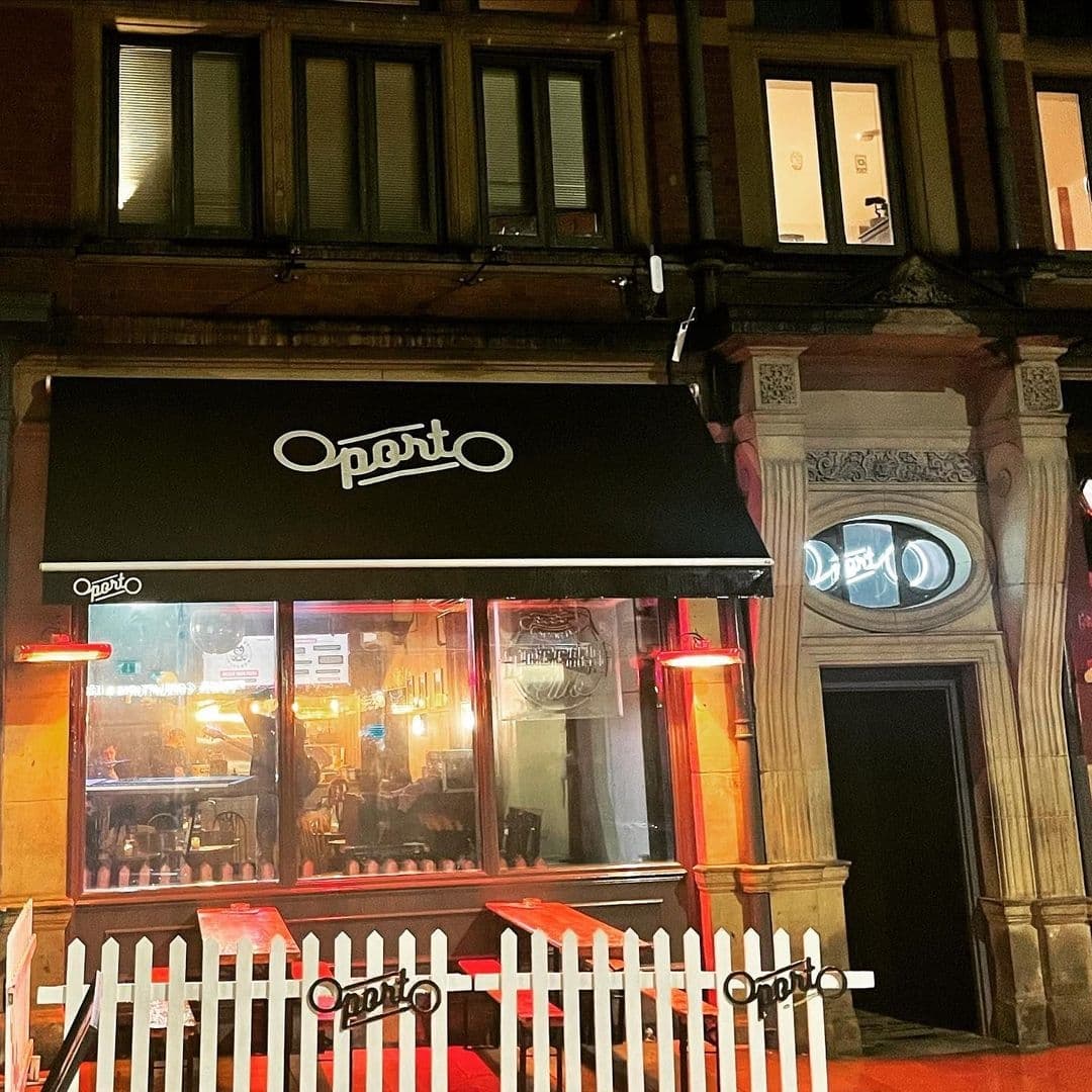 Night view of a lively Oporto restaurant with illuminated signage, patrons visible inside, and a white picket fence in front, set against a traditional building facade in Oporto.