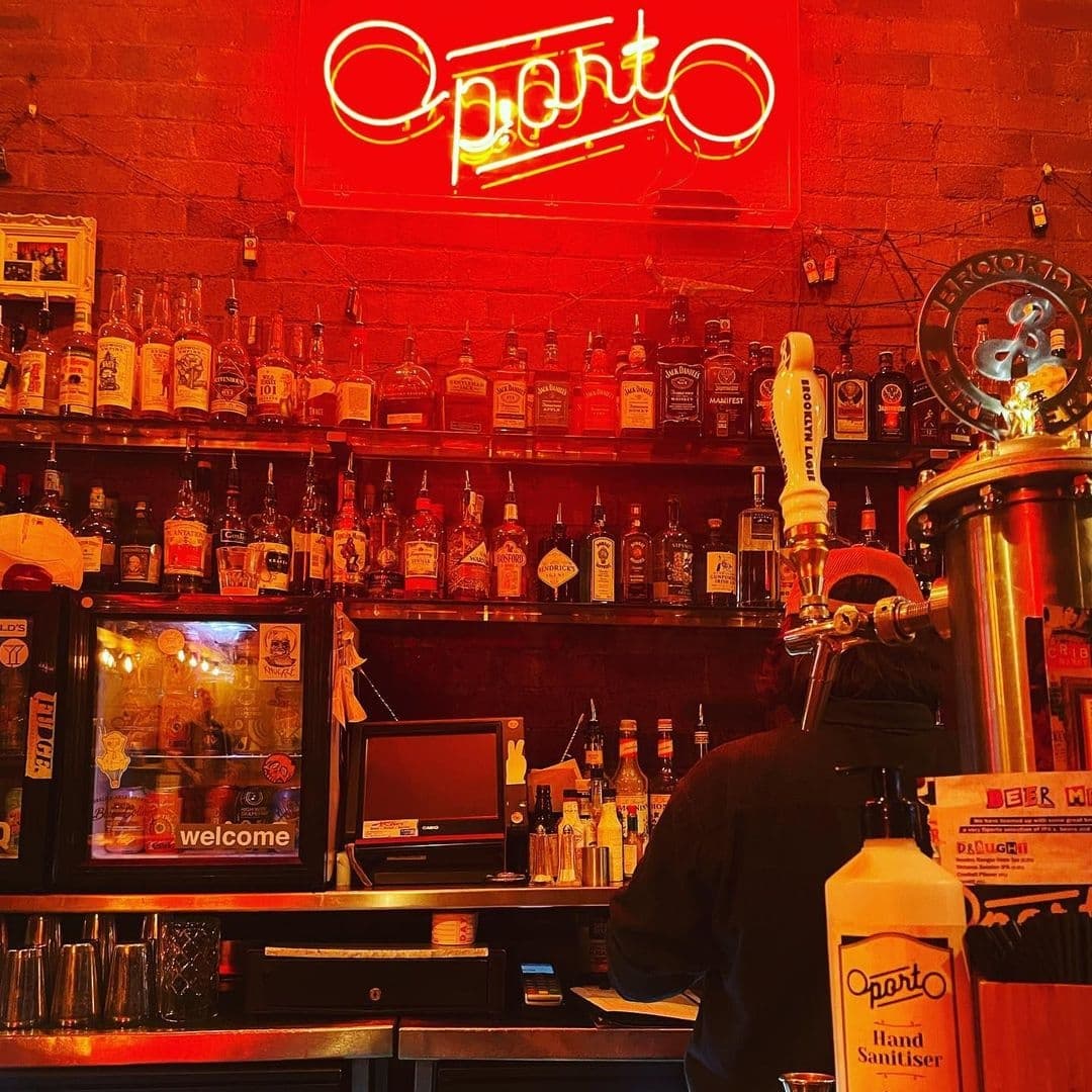 "Cozy bar ambiance at Oporto with neon sign, shelves of whiskey bottles, beer taps, and bartender at work."