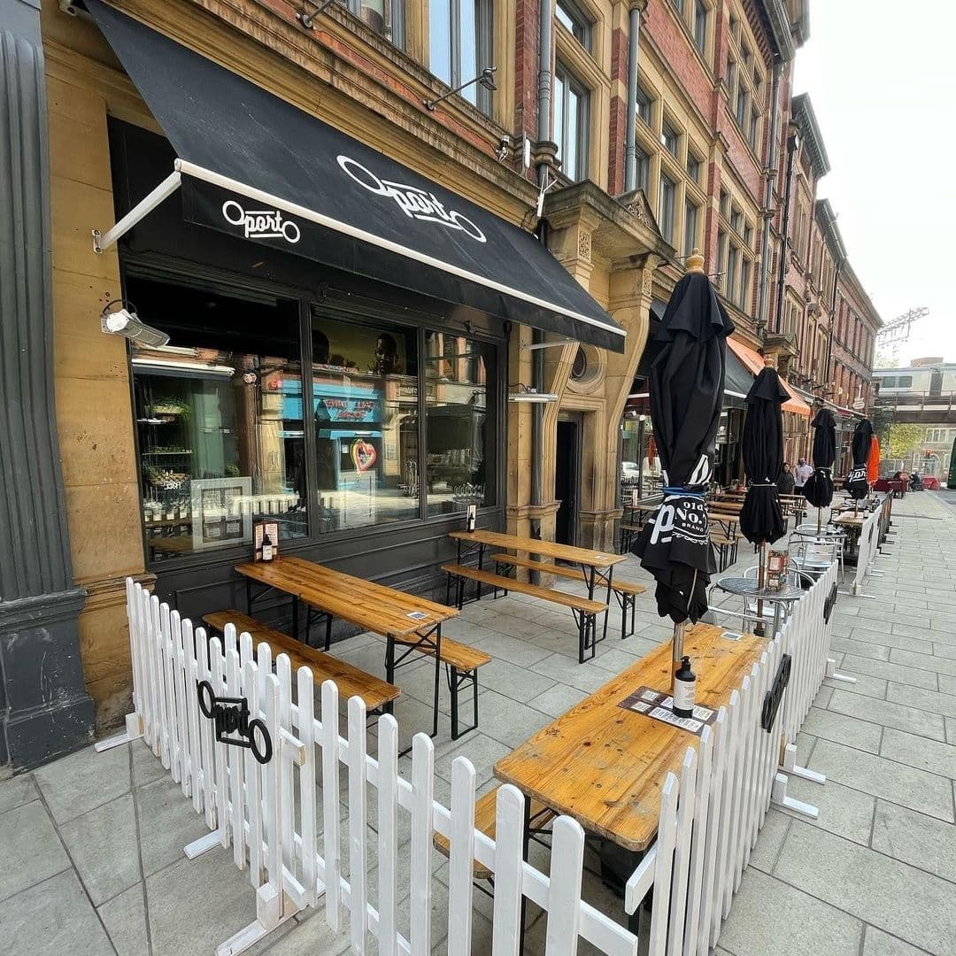 Outdoor seating at Oporto restaurant with wooden tables and benches, white picket fence, and folded black umbrellas on a city sidewalk.