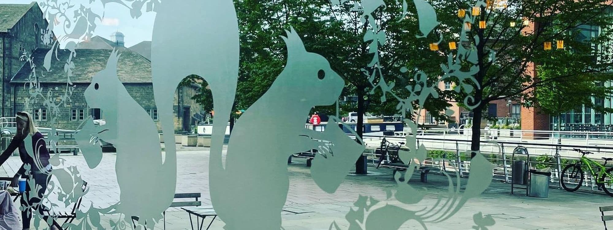 Artistic frosted glass panel with woodland creatures at Out of the Woods, overlooking an urban plaza with buildings, trees, and passersby.