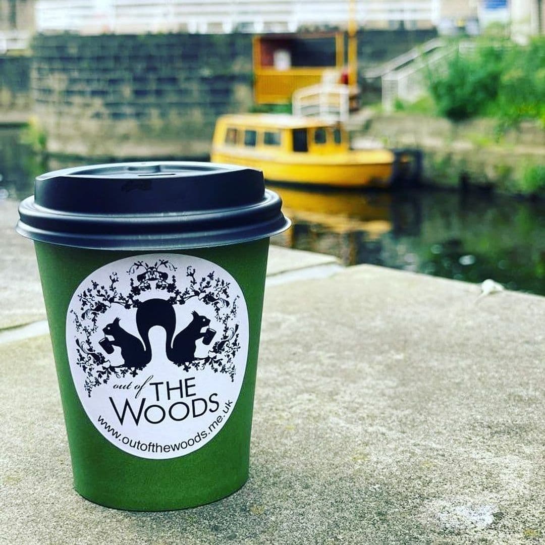 "Green takeaway coffee cup with 'Out of the Woods' logo in focus, placed on a stone surface with a blurred background featuring a yellow boat on water."