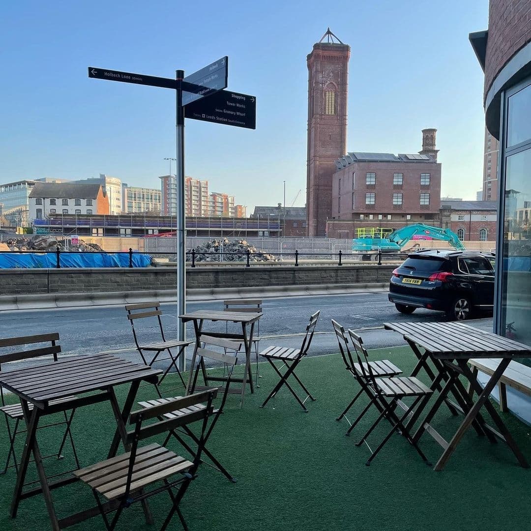 Outdoor seating area at "Out of the Woods" with wooden tables and chairs on green artificial turf, directional signpost in the foreground, and historic red brick tower in the background under a clear blue sky.