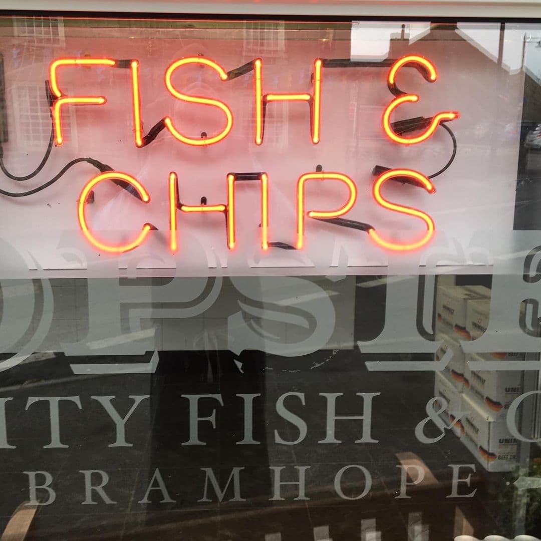 Neon sign reading 'Fish & Chips' at Popsies of Bramhope, with reflection of street visible in window.