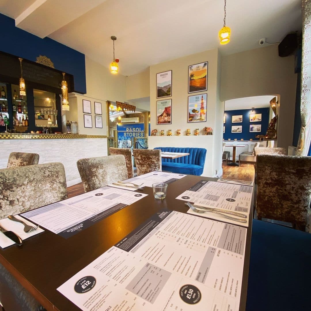 Interior of Rasoi Stories restaurant showing a cozy dining area with menus on tables, plush seating, and a bar in the background. Decor includes hanging pendant lights and framed pictures on the walls.