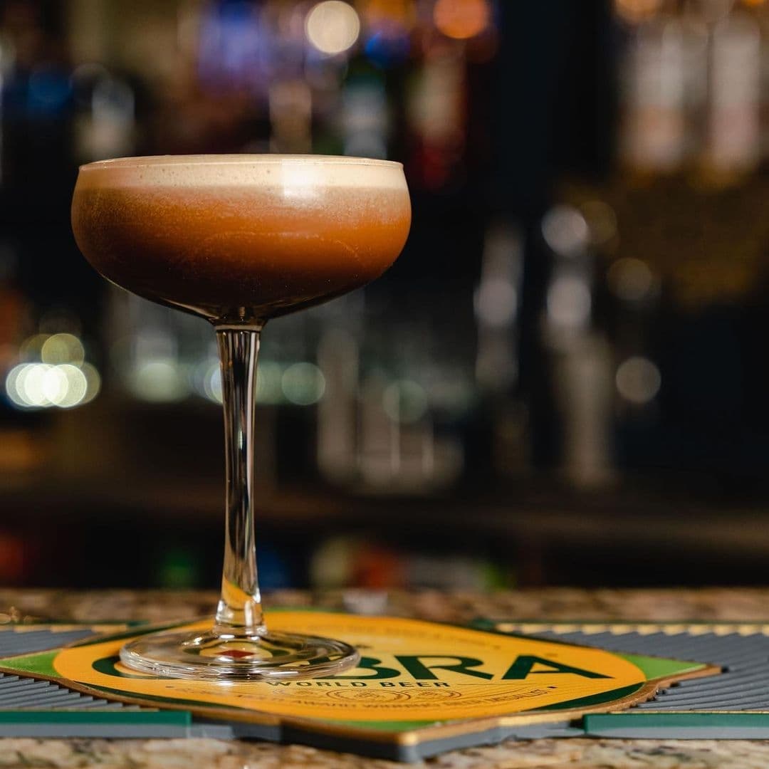 Craft cocktail on a bar counter at Rasoi Stories, with a frothy top and a warm amber color, presented in an elegant stemmed glass, with a softly blurred background of a bar setting.