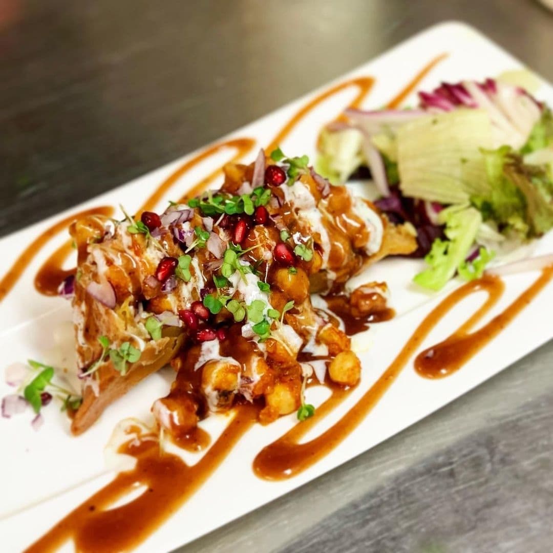 Gourmet dish served at Rasoi Stories, featuring crispy golden-brown pieces garnished with pomegranate seeds, fresh herbs, diced onions, and drizzled with a rich brown sauce, accompanied by a side salad.