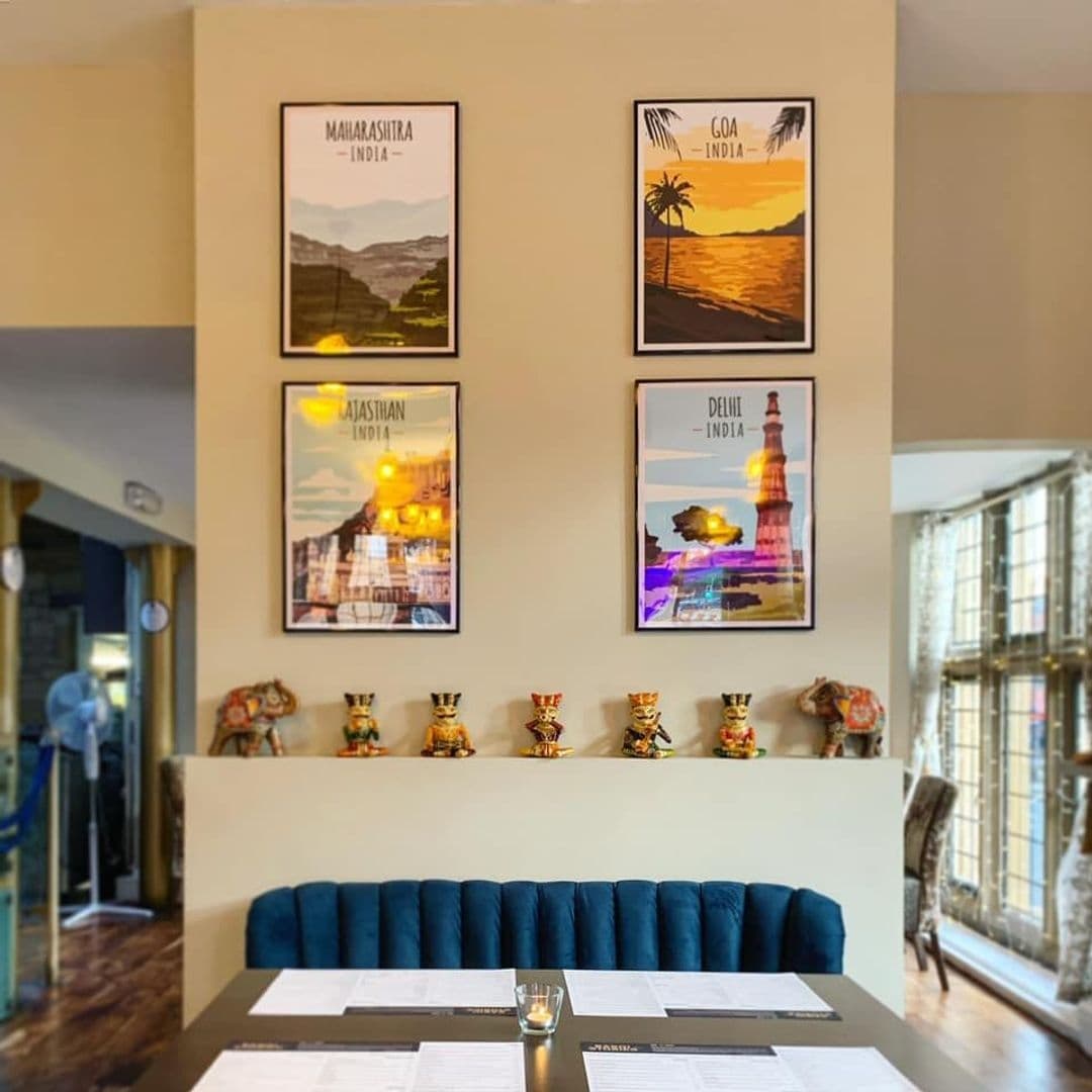 Interior of Rasoi Stories with a cozy dining setup featuring a blue upholstered bench, neatly arranged tables with menus, and decorative Indian-themed artwork on the wall.