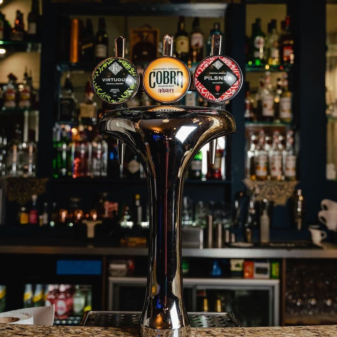 Shiny beer taps with Cobra and Pilsner labels at Rasoi Stories bar, with a selection of spirits and beverages in the background.