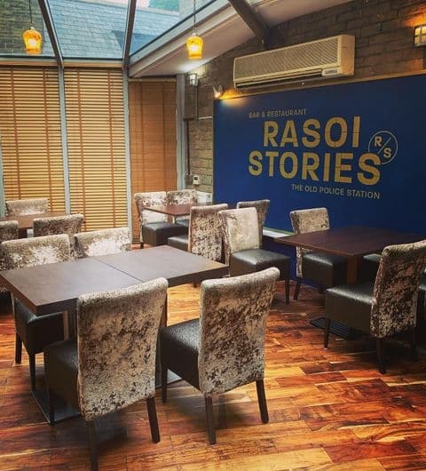 Interior of Rasoi Stories restaurant showing dining tables and chairs with a blue wall featuring the restaurant's logo and the text 'The Old Police Station'.