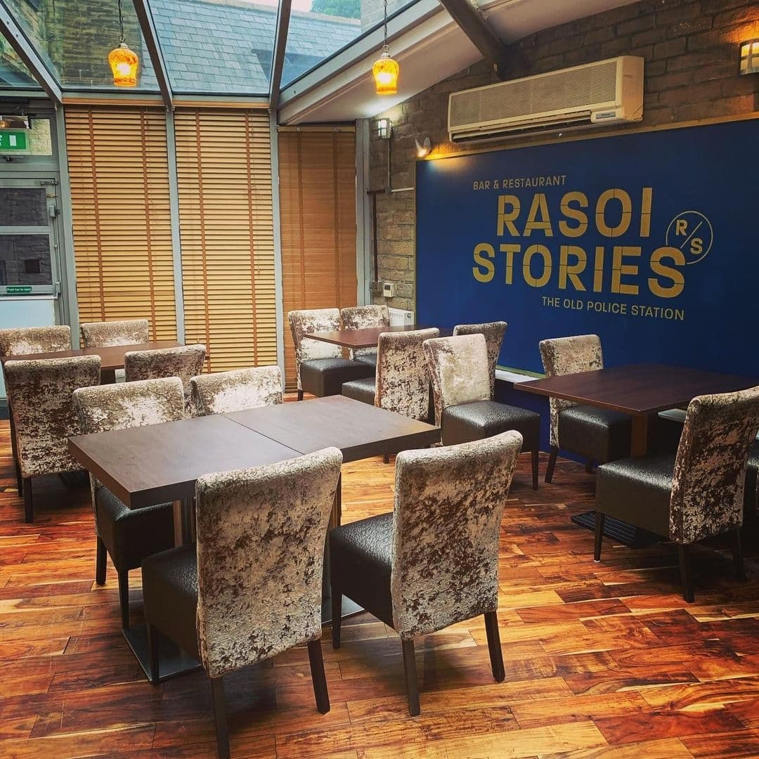 Interior of Rasoi Stories restaurant with elegant dining tables and plush chairs, warm lighting, and a prominent blue wall with the establishment's logo.