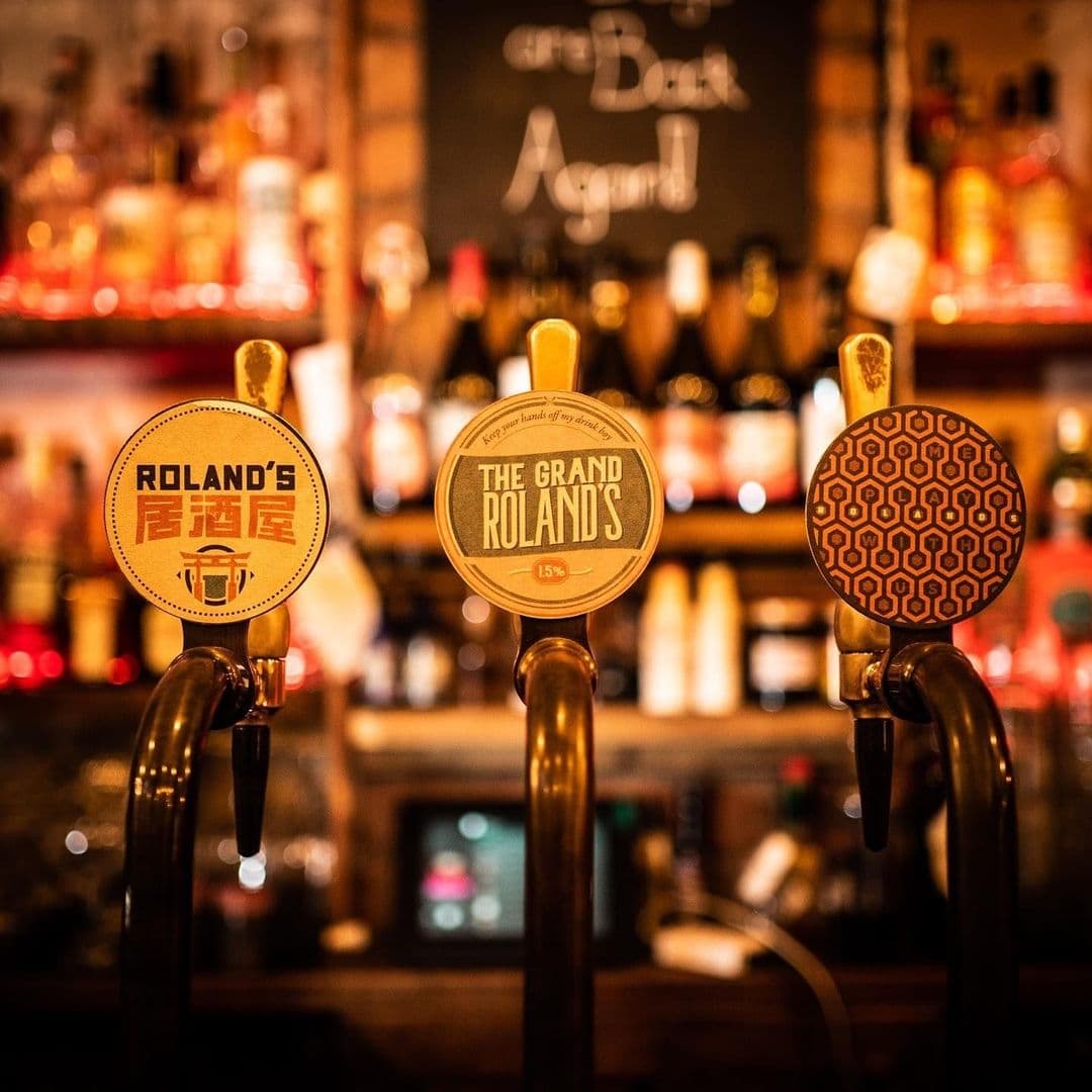 Close-up of three beer tap handles at Roland's bar, with the first and second featuring the Roland's logo and the third with a geometric pattern.