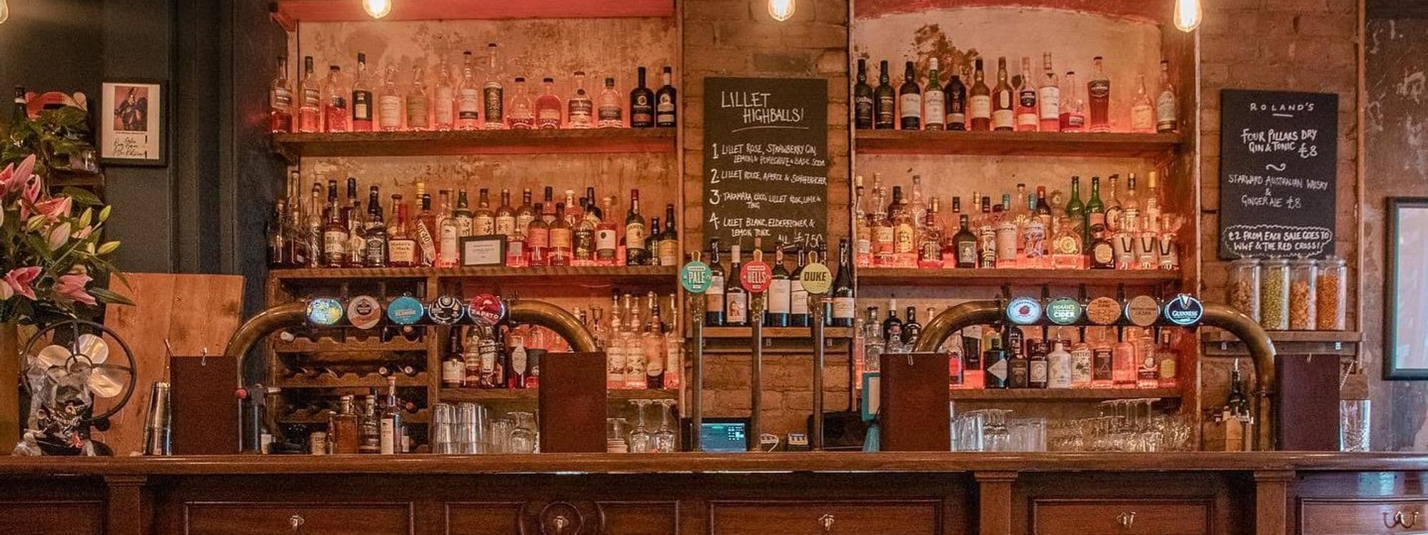 Interior view of Roland's bar with a wooden counter, a variety of spirits on shelves, beer taps, and a chalkboard menu displaying drink options and prices.