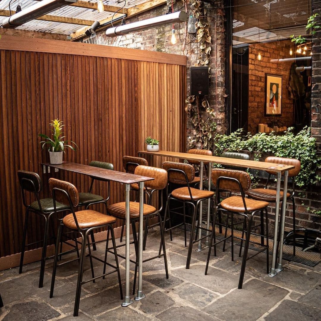 Cozy outdoor seating area at Roland's with wooden high-top tables, brown leather bar stools, and lush greenery on a brick backdrop.