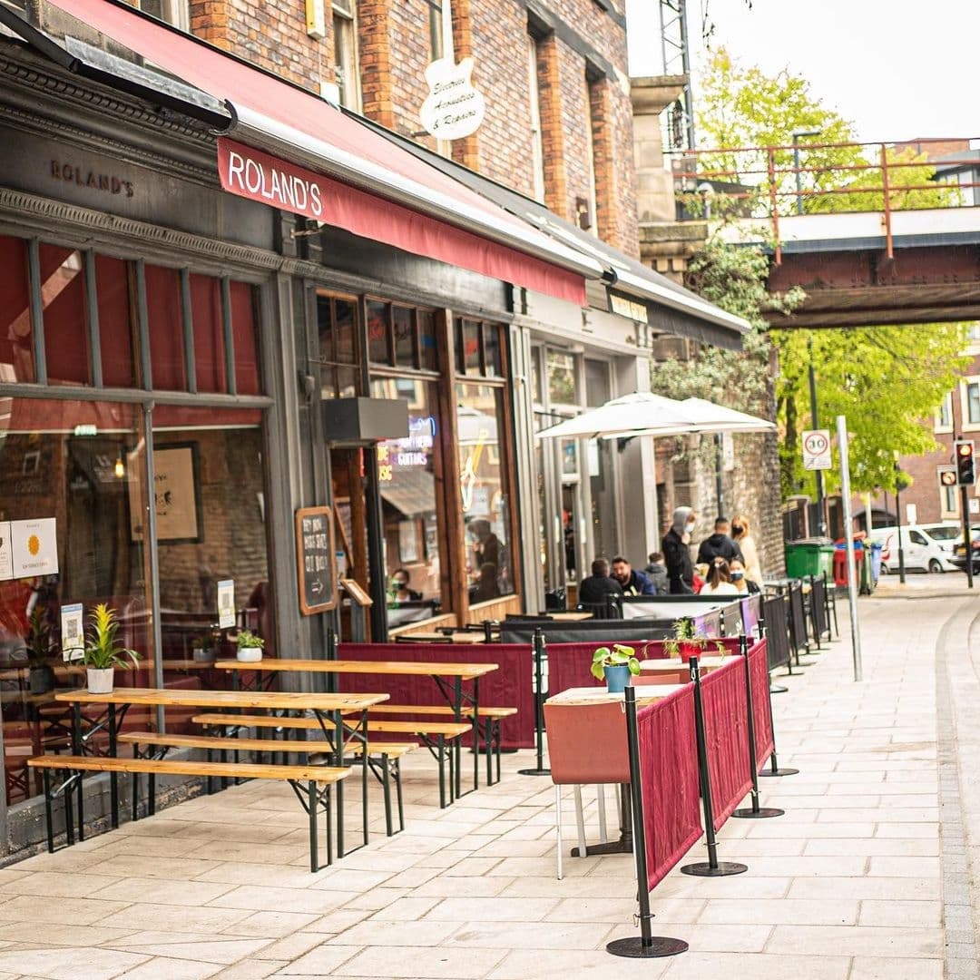 Exterior view of Roland's with outdoor seating and red barriers along a cobbled street, patrons enjoying drinks under an umbrella, and a warm, inviting atmosphere.