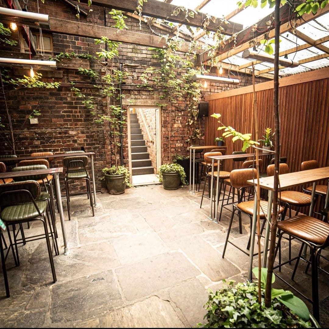 Cozy outdoor patio at Roland's with rustic brick walls, green climbing plants, wooden tables, metal chairs, and a glass roof partially covered by foliage.