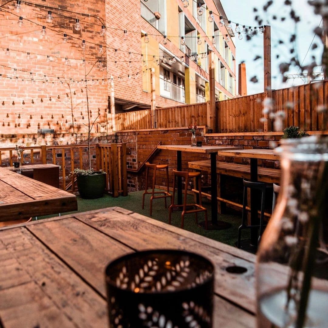 Cozy outdoor seating at Roland's with wooden tables and benches, string lights overhead, and a warm brick wall backdrop.