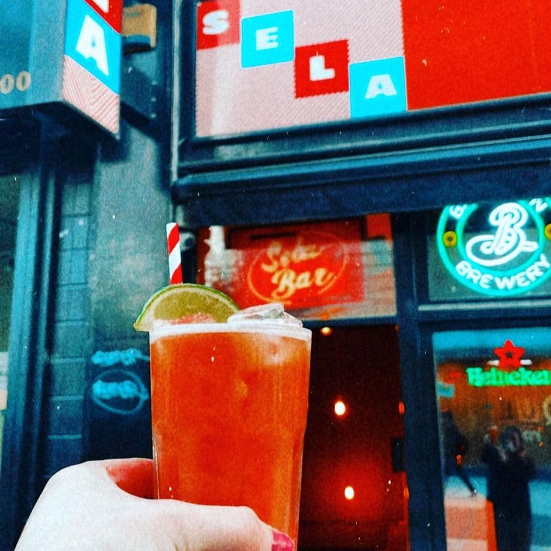 Close-up of a hand holding a cocktail with a lime slice and striped straw in front of Sela Bar, with the bar's sign and entrance visible in the background.