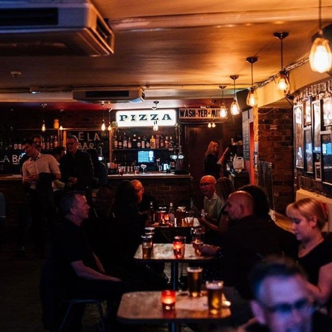 Patrons enjoying a cozy evening at Sela Bar, an ambient venue with dim lighting, exposed brick walls, and a bar serving pizza.