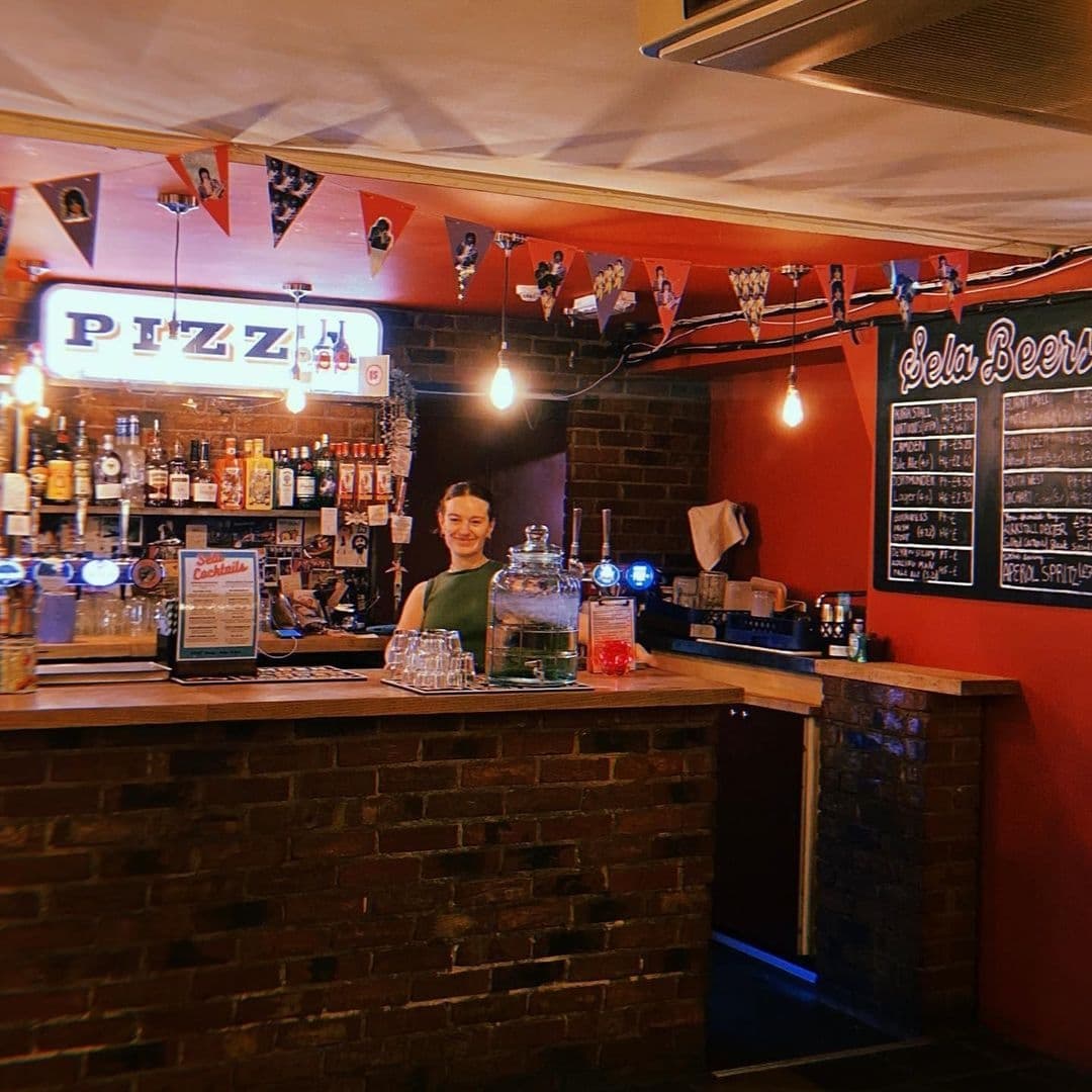 Friendly bartender standing behind the counter at Sela Bar with a selection of drinks, under a neon pizza sign and festive bunting.