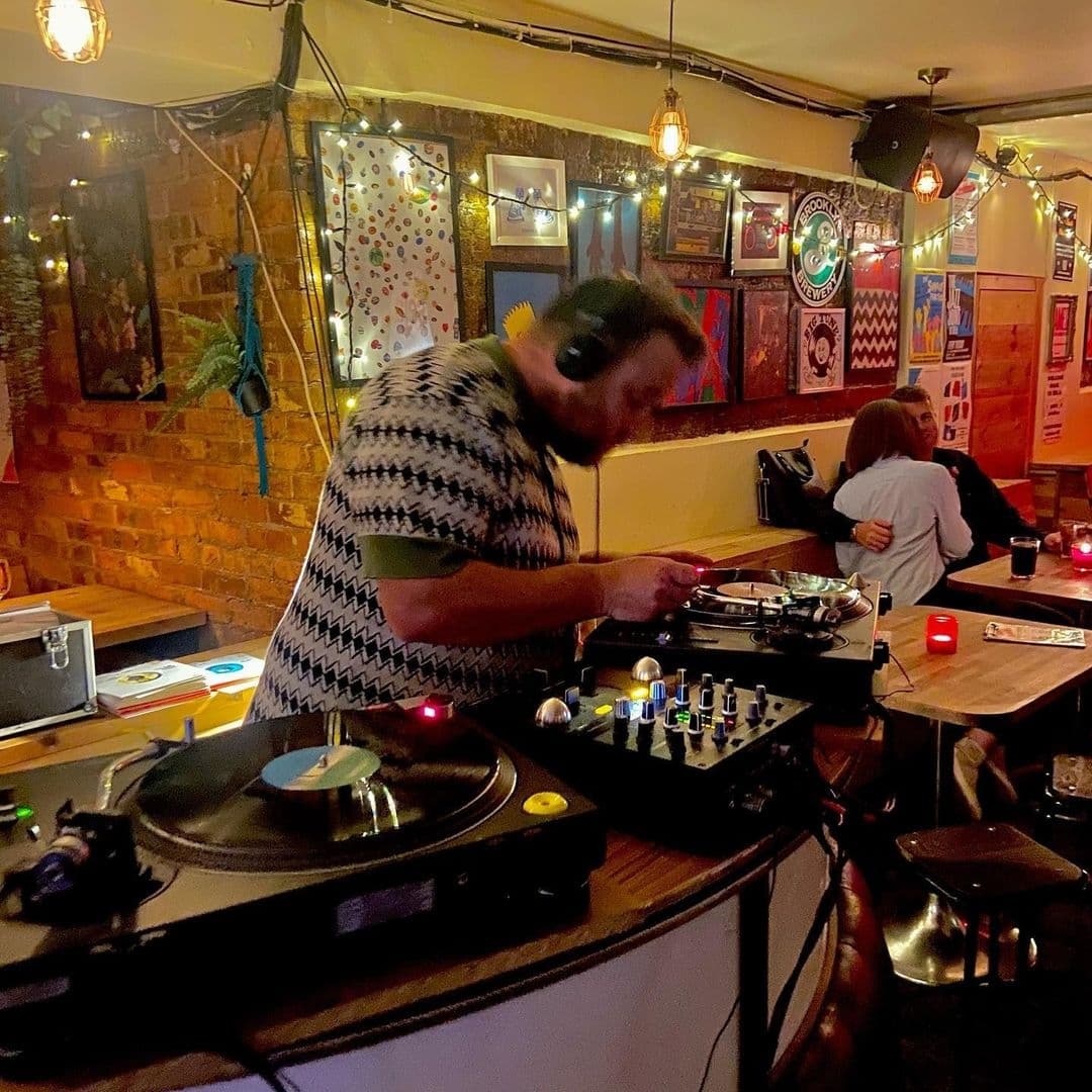 DJ playing vinyl records at Sela Bar with patrons in the background enjoying drinks and conversation.