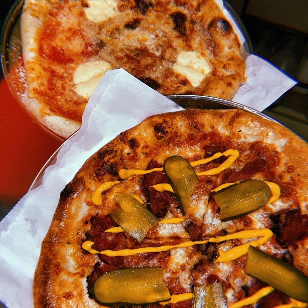 Two freshly baked pizzas at Sela Bar, one topped with pickles and drizzled with mustard, the other with a simple cheese topping.