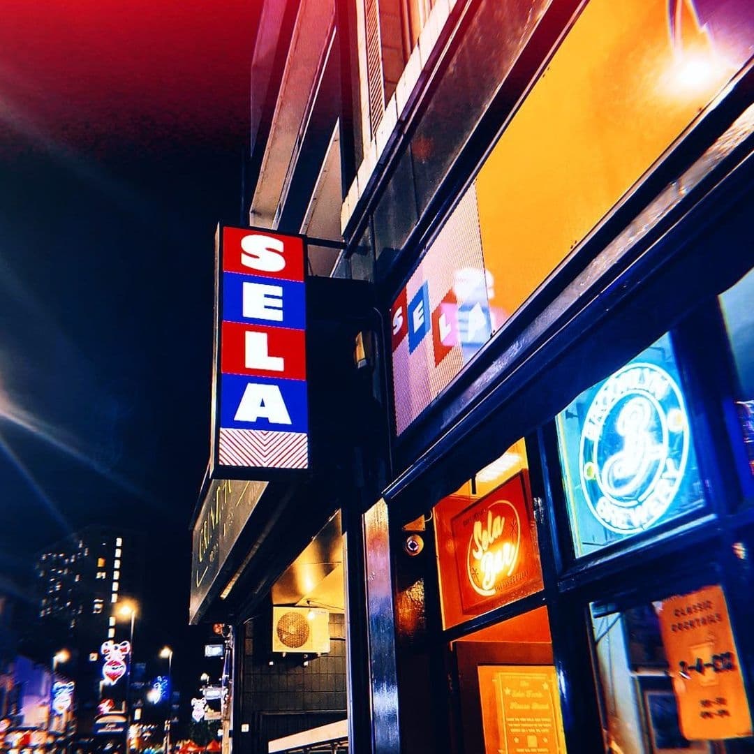Exterior view at night of Sela Bar with illuminated signage and colorful facade.