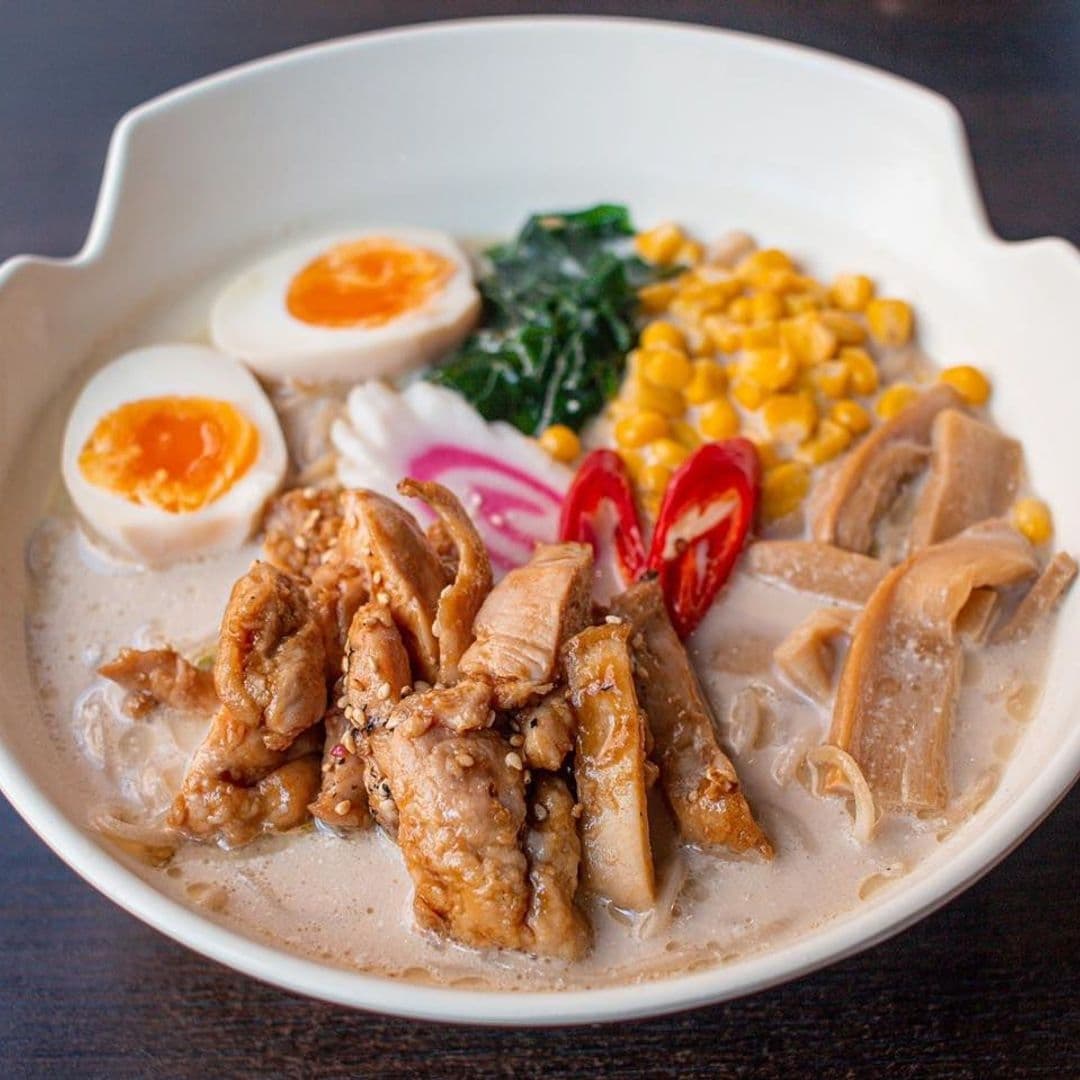 Alt text: "A bowl of traditional ramen from Sushi Waka, featuring sliced chicken, soft-boiled egg, corn, bamboo shoots, and greens, garnished with red chili peppers."