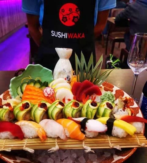 Assorted sushi and sashimi platter at Sushi Waka, with a server wearing a branded apron in the background.