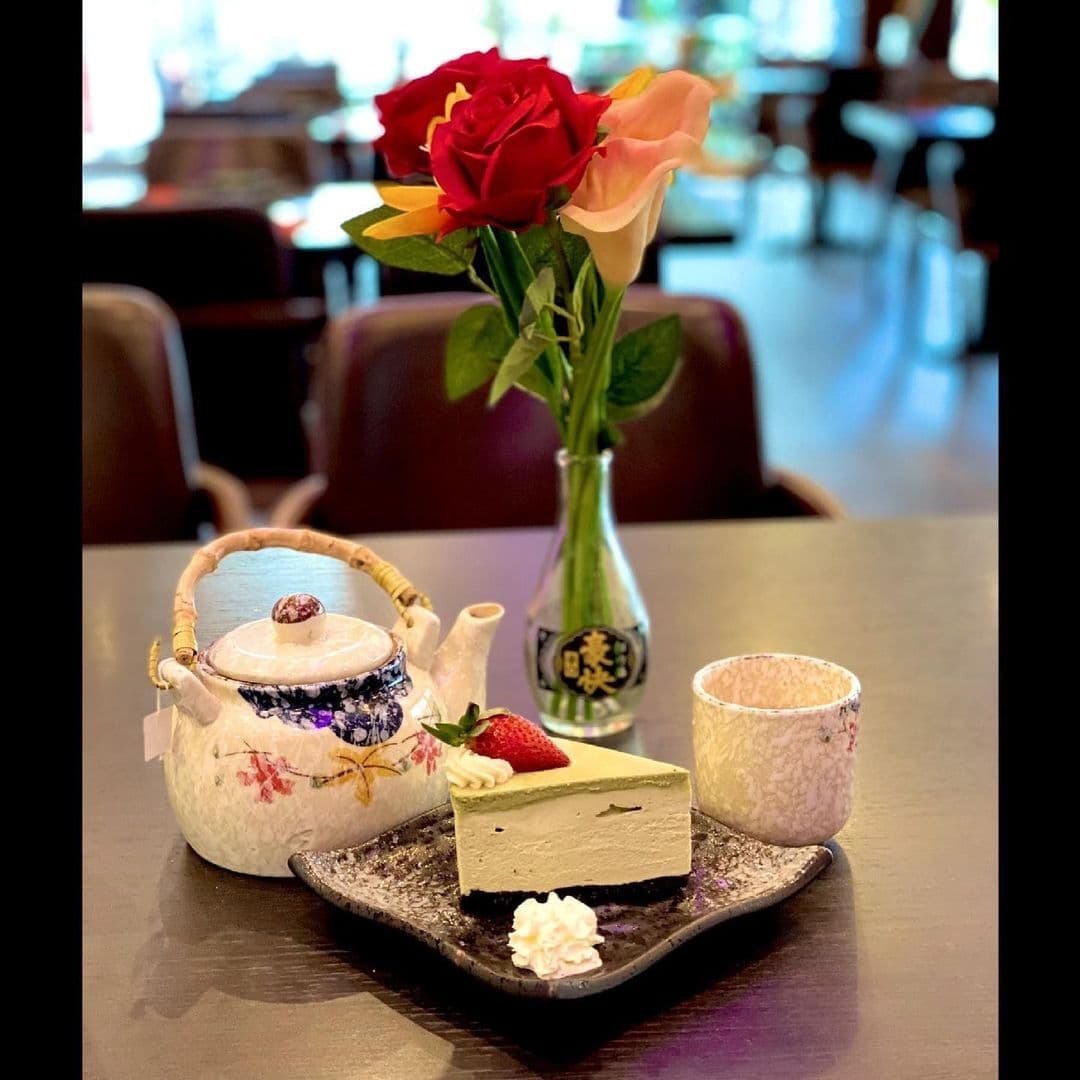 Alt text: "Elegant dining setup at Sushi Waka featuring a traditional Japanese teapot and cup, with a slice of matcha cheesecake garnished with a strawberry and cream, presented on a black plate. A vase with a red and a peach rose adds a touch of natural beauty to the experience."
