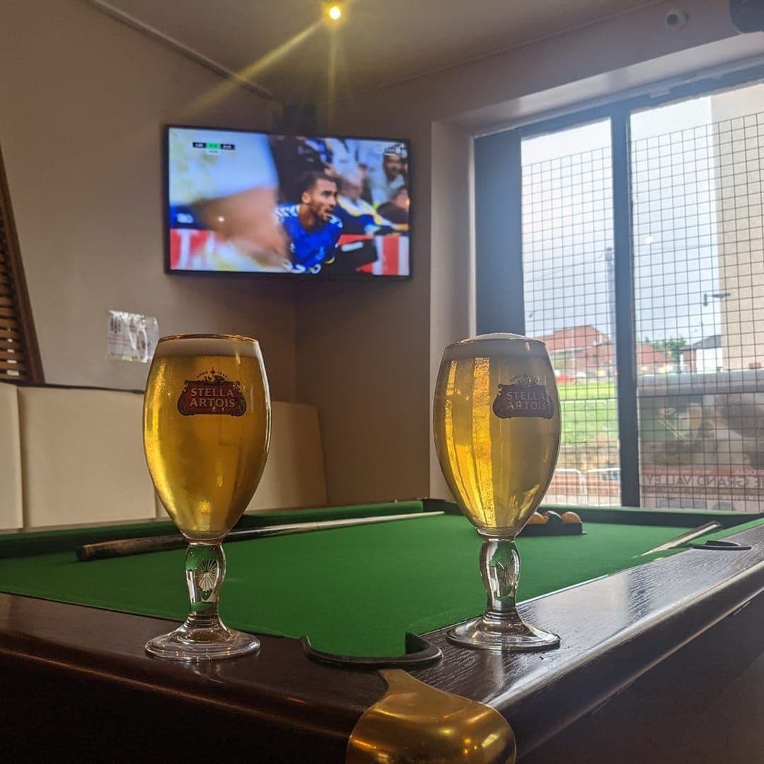 Two glasses of Stella Artois beer on a pool table at The Grand Valley Bar & Kitchen with a sports game broadcasting on a television in the background.