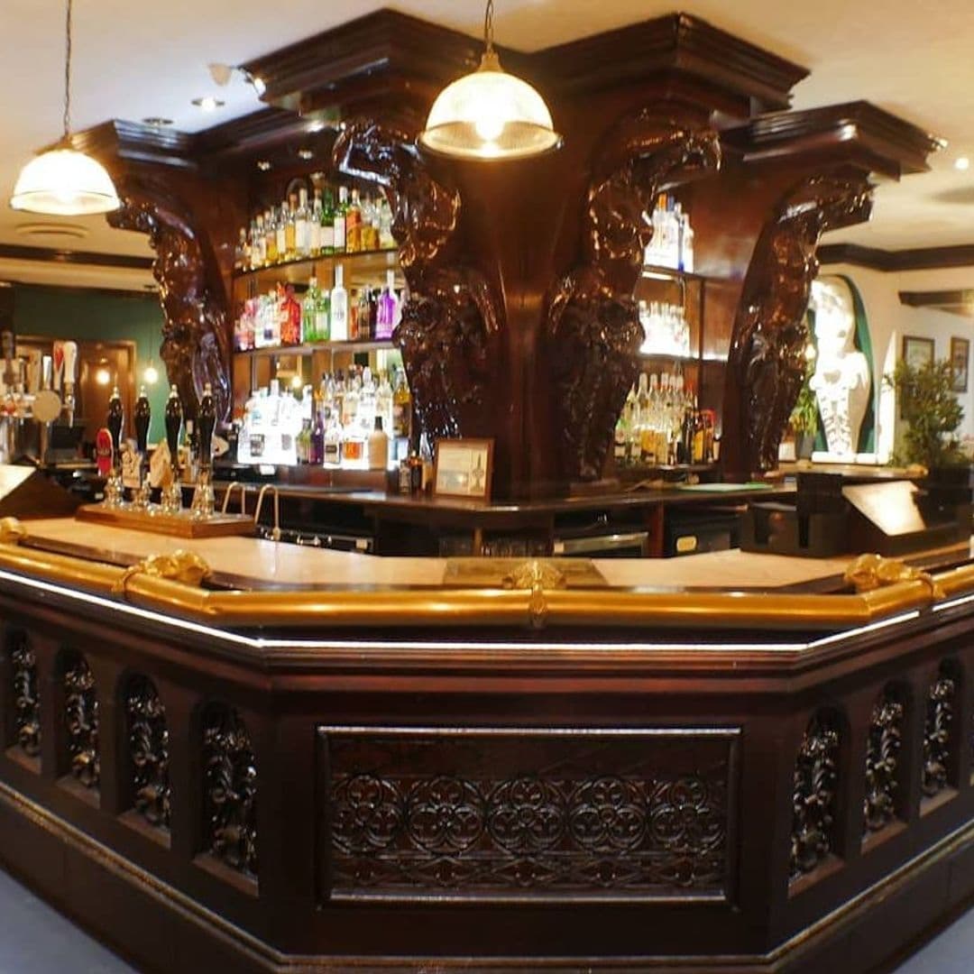 Elegant wooden bar counter with ornate carvings at The New Conservatory, featuring a selection of spirits on shelves and pendant lighting above.
