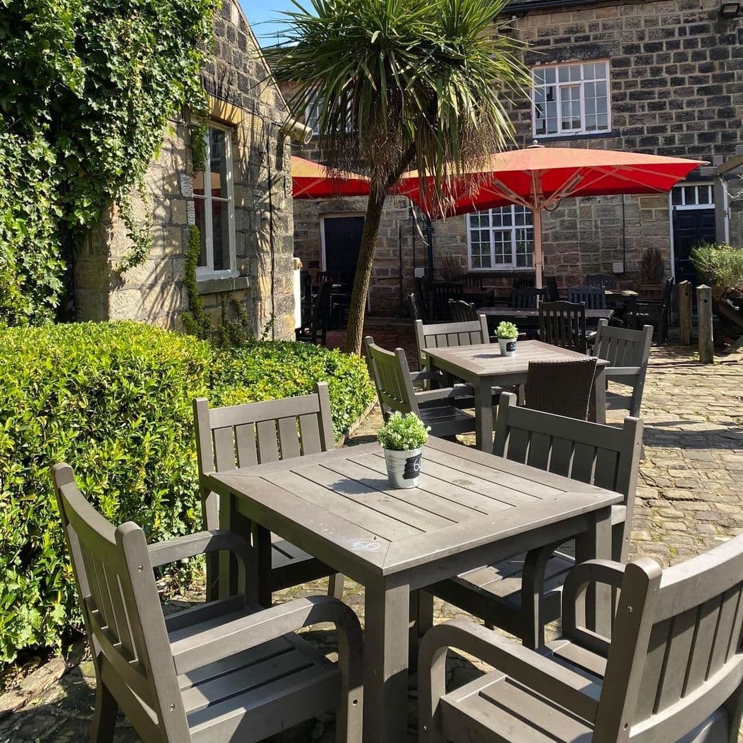 Outdoor patio area at The Roundhay Fox with wooden tables and chairs, red umbrellas, surrounded by greenery and a stone building in the background.