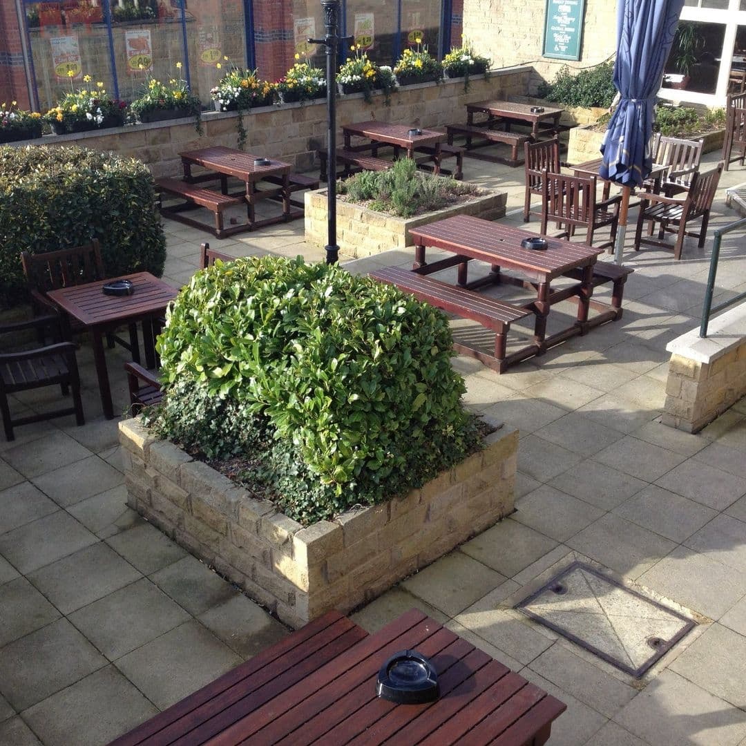 Outdoor seating area at The Three Hulats with wooden benches and tables, surrounded by green shrubs and a clear blue sky.
