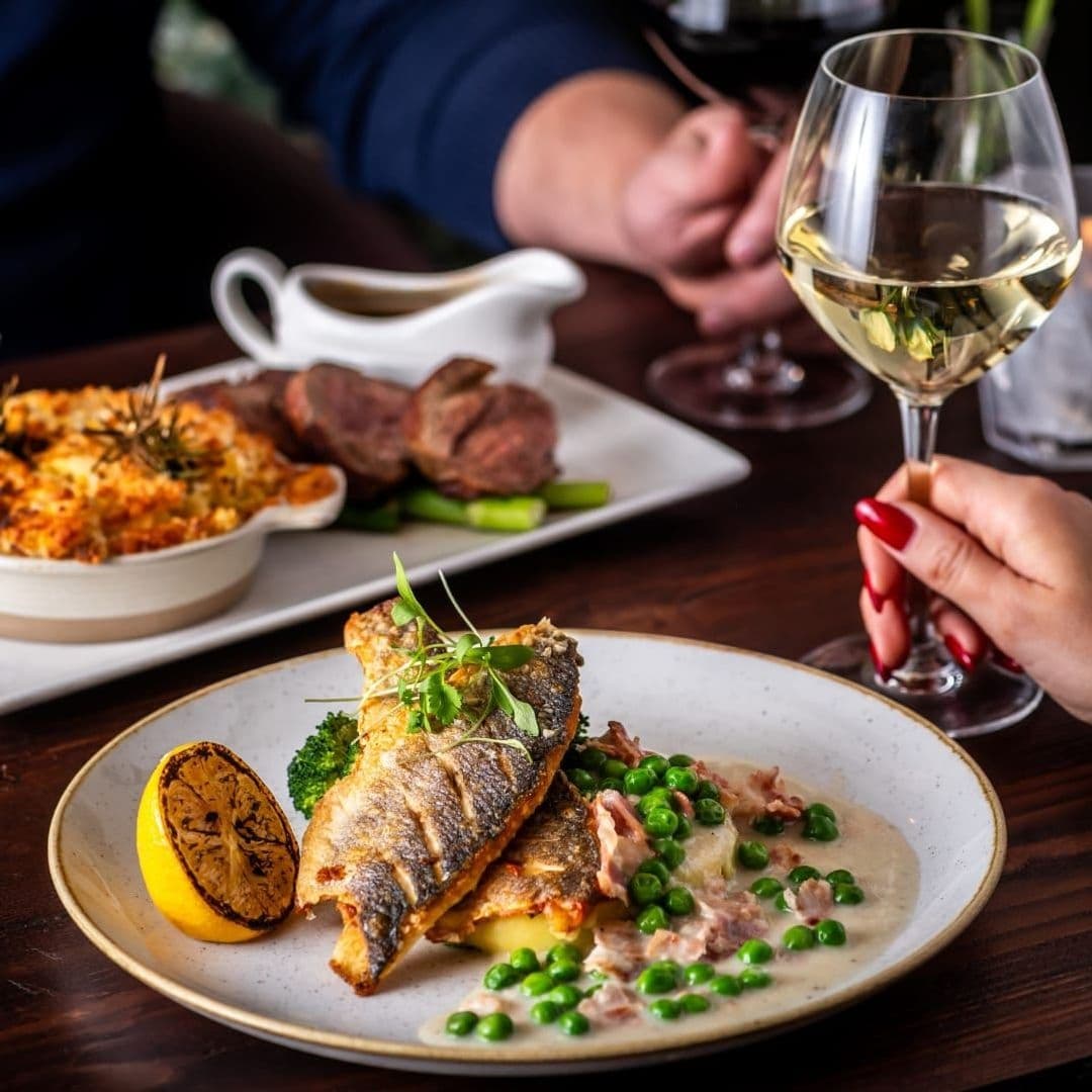 Gourmet dining experience at The Roundhay Fox, featuring a plate of grilled fish with peas and a lemon wedge, accompanied by a glass of white wine and a dish of steak and mashed potatoes in the background.