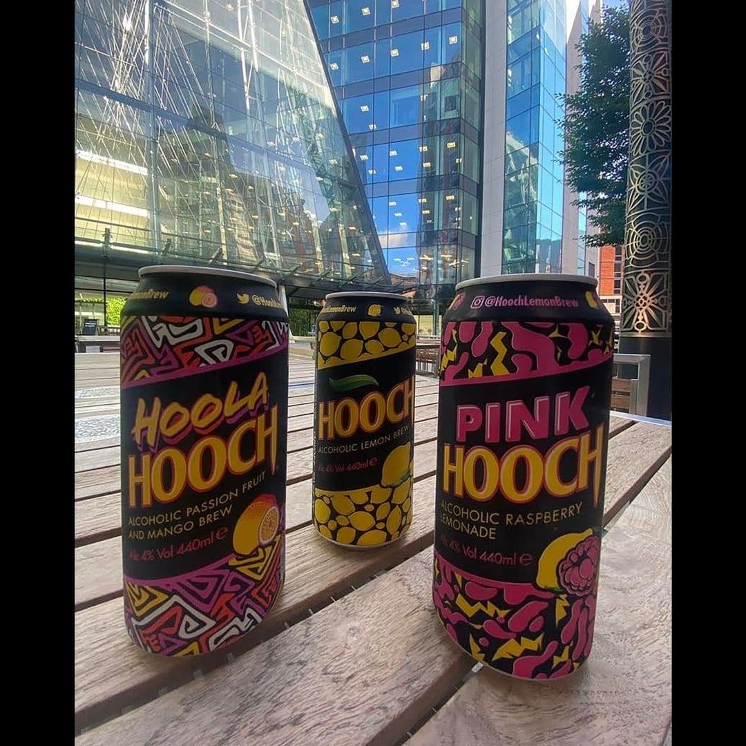 Three colorful cans of Hooch alcoholic beverages on a wooden table at Toast Bar, with a modern glass building in the background.