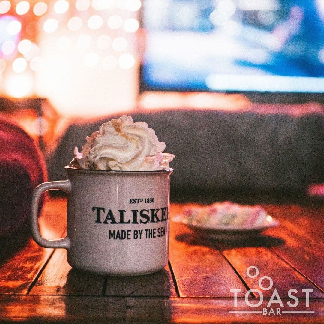 A cozy mug topped with whipped cream on a wooden table at Toast Bar, with warm lighting and bokeh in the background.